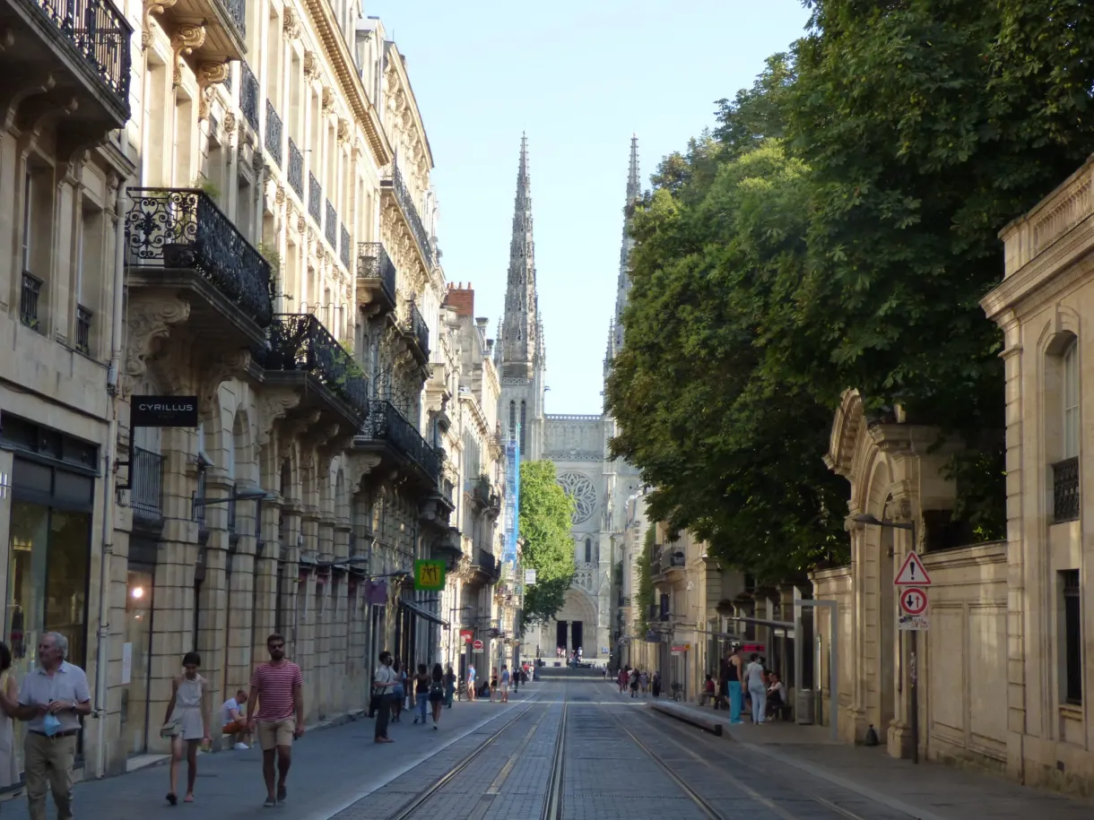 Rue commerçante ombragée où passent des promeneurs, flèche en perspective