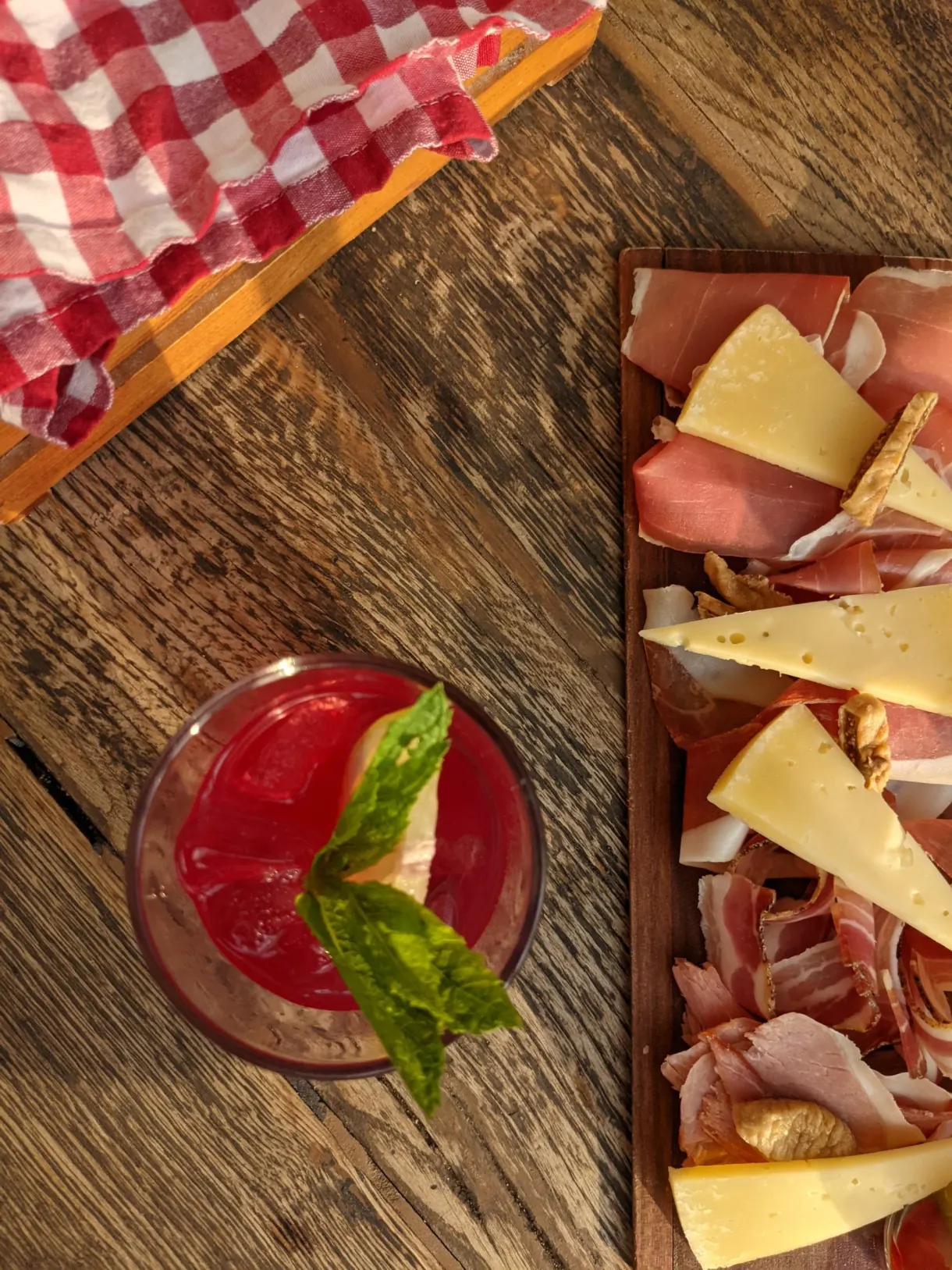 Board of cured meats and cheeses with a red cocktail on raw wood