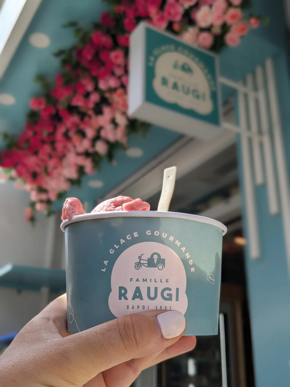 Tub of ice cream held in front of a flower-decked turquoise shopfront