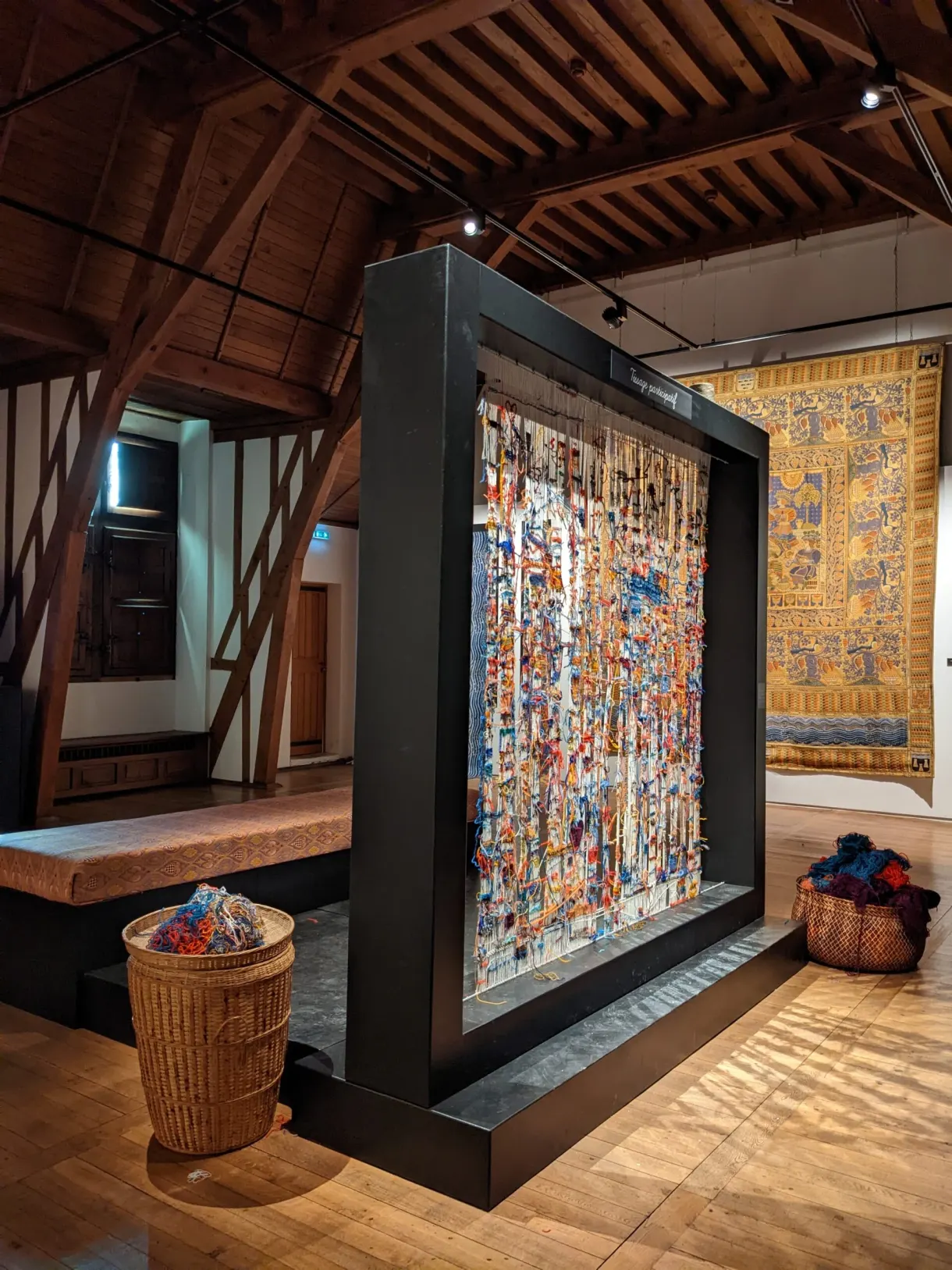 Large colourful tapestry displayed beneath a wooden roof frame