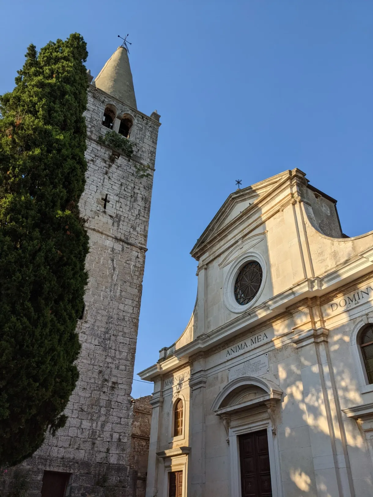 Bell tower and pale façade of the church of Bale behind a tall cypress